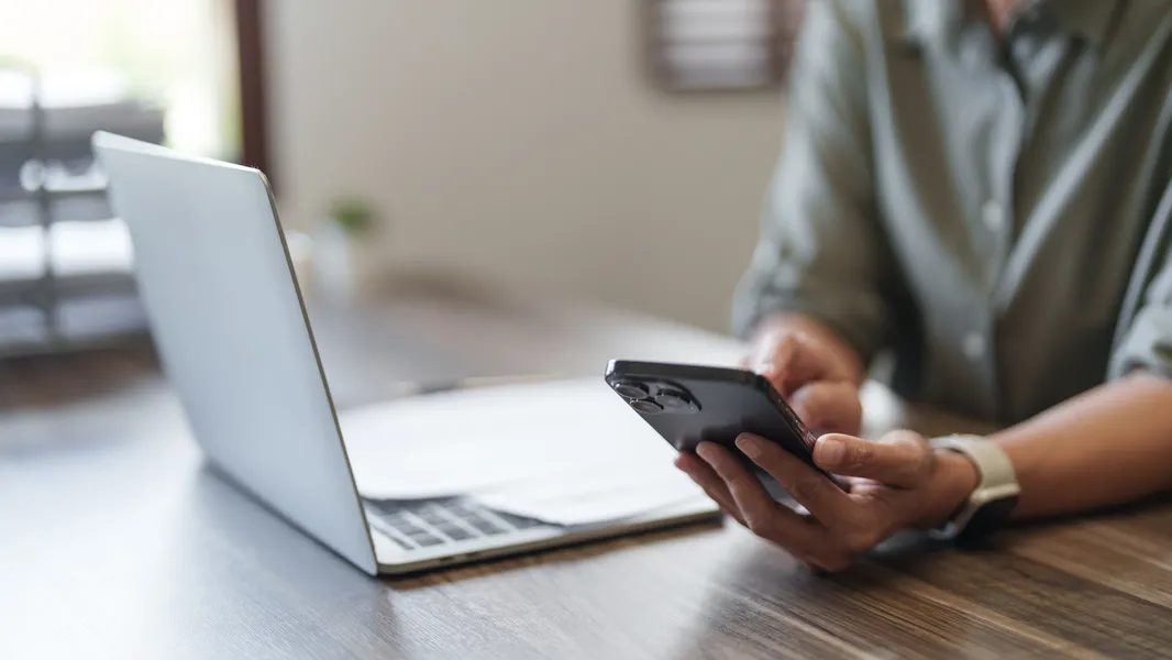 A person's hands using their mobile phone while sitting in front of a laptop